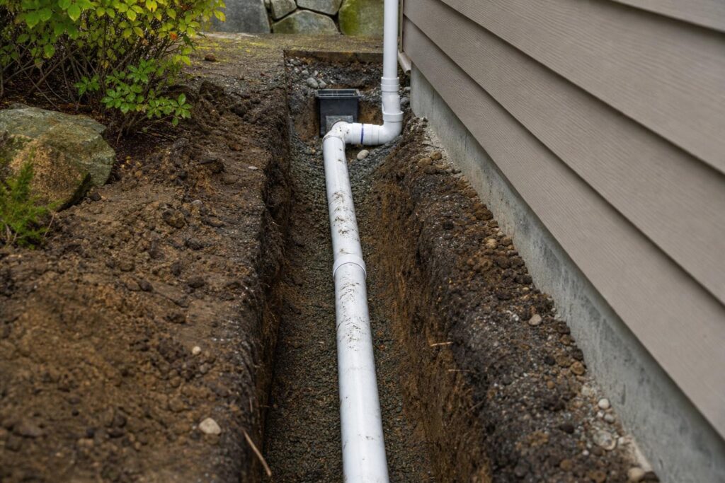 White PVC drainage pipe installed in a deep dirt trench alongside a residential house foundation for professional exterior water management.