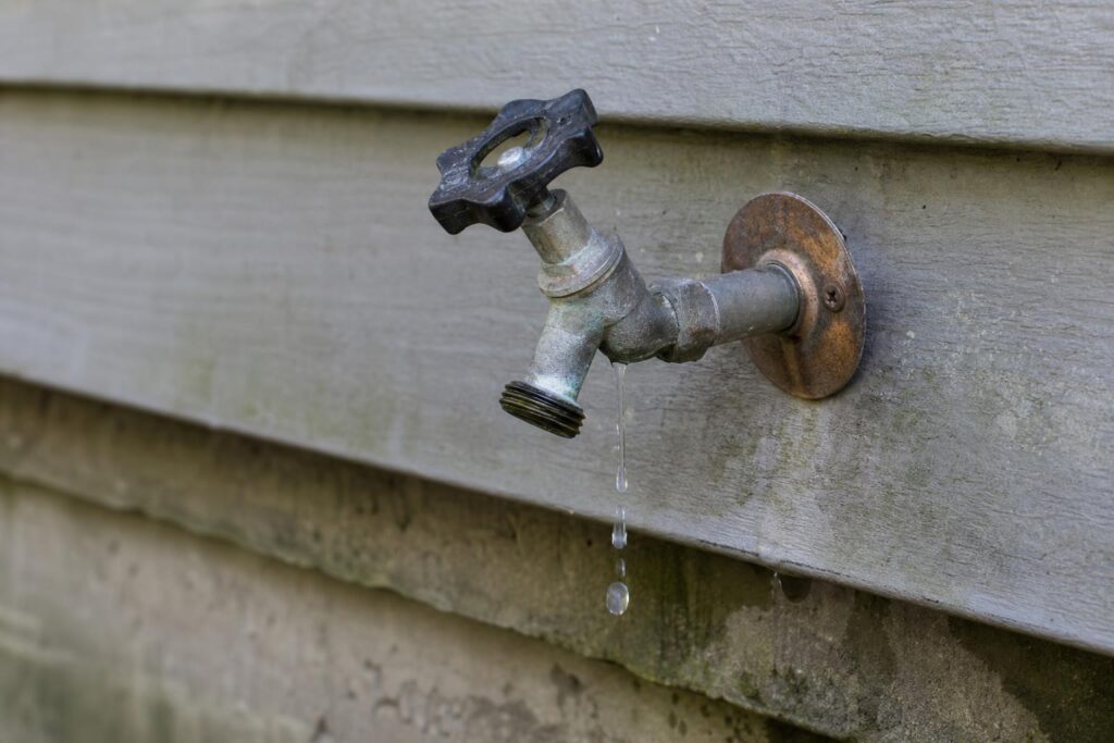 Weathered metal outdoor faucet with a black handle dripping water onto a grey wooden house siding.