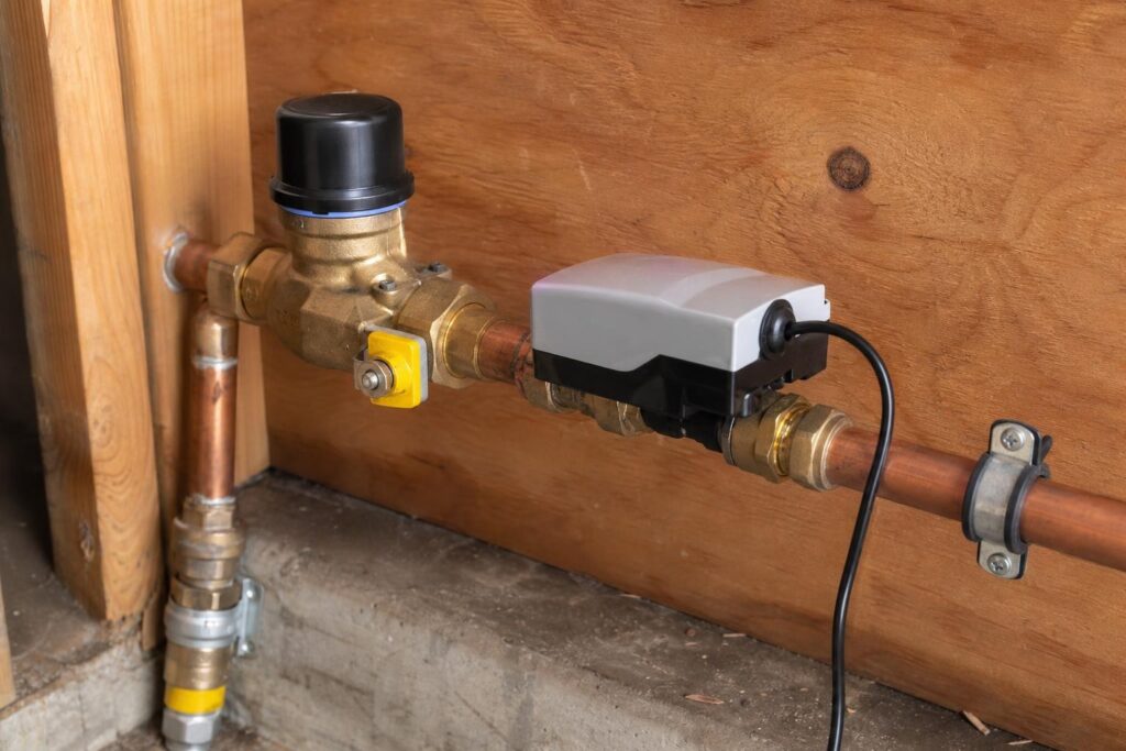 Automatic water shut-off valve with a grey electronic controller installed on a copper main water line against a plywood wall.